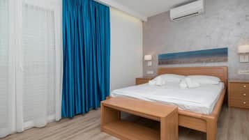 A hotel room with a bed, a bedside table, a bench, an air conditioning unit, and a painting on the wall.