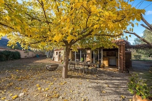A patio area with a table and chairs under a tree with yellow leaves, a small brick structure, and a gravel ground.