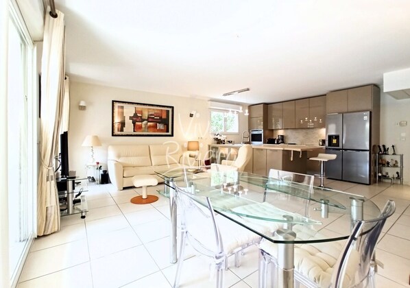 A modern living room with a glass dining table, a sofa, and a kitchen area with stainless steel appliances.