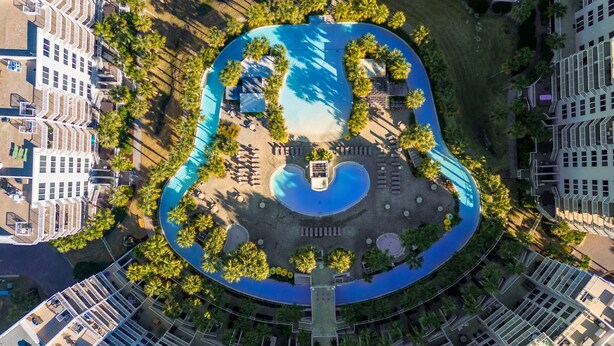 Ett flygbildsfoto av ett hotell med en central pool som är formad som en gitarr, omgiven av palmer och avslappningsområden.