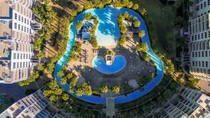An aerial view of a hotel with a central swimming pool shaped like a guitar, surrounded by palm trees and lounge areas.