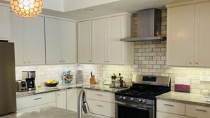 Una cocina moderna con armarios blancos, un lavamanos de acero inoxidable y una estufa a gas. Hay dos lámparas colgantes con pantallas hexagonales.