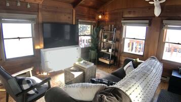 A cozy cabin interior with a sofa, chairs, a TV, wooden walls, and a deer head decoration.