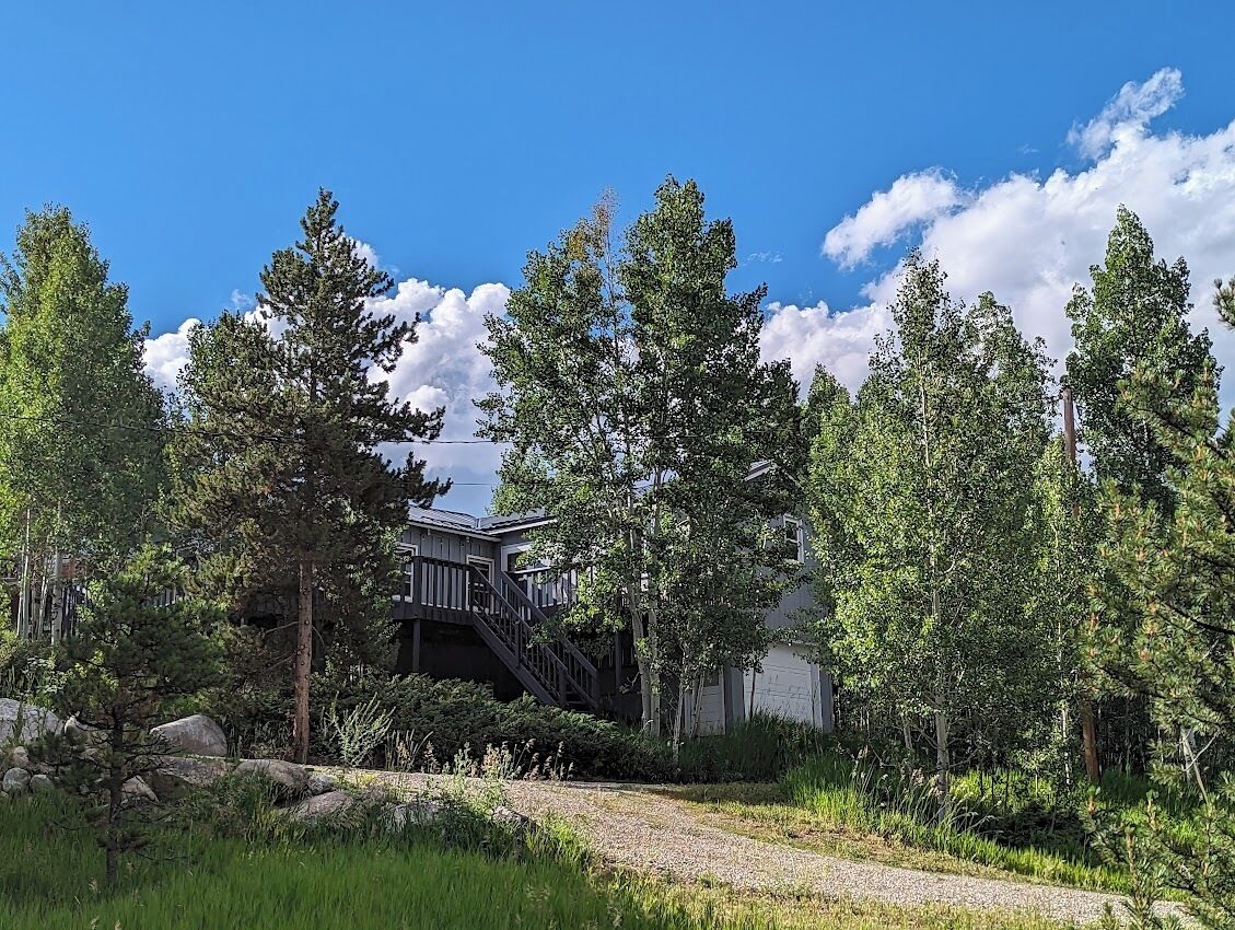 A house with a wooden deck and stairs leading to a garage, surrounded by tall trees and a gravel path.