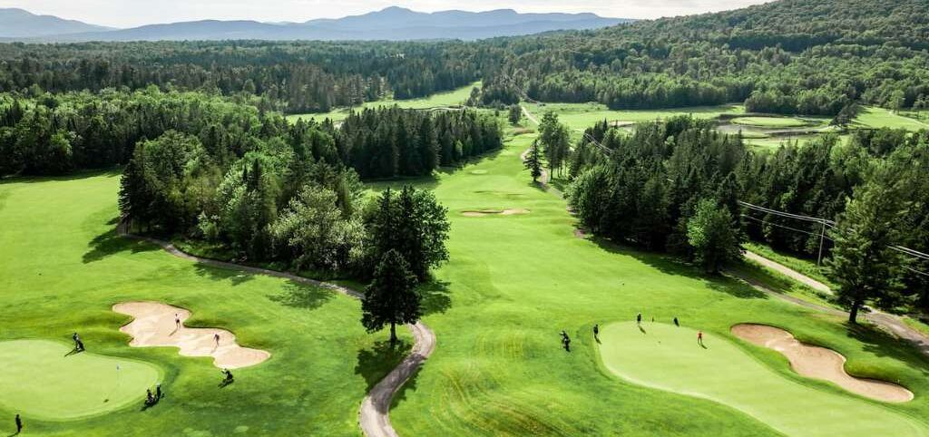 Vue aérienne d’un parcours de golf comportant plusieurs trous, entouré de forêts denses et bordé de montagnes lointaines.