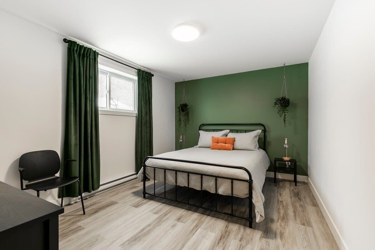 Une chambre à coucher avec un lit, un bureau et une chaise. On y trouve deux plantes suspendues et une fenêtre avec des rideaux.