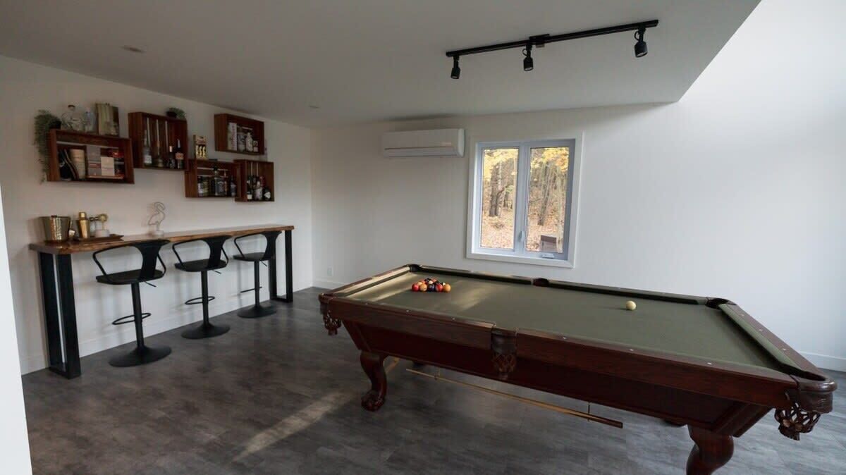 Una mesa de billar con sillas, una zona de bar con taburetes y una ventana que da a vistas de árboles.