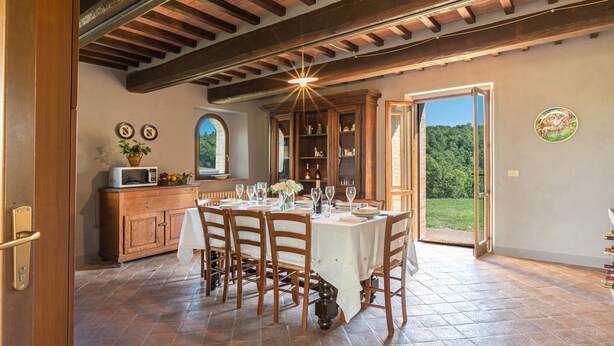 A dining area with a table set for four, wooden chairs, a wooden cabinet with a microwave, and a view of the outdoors through a door.
