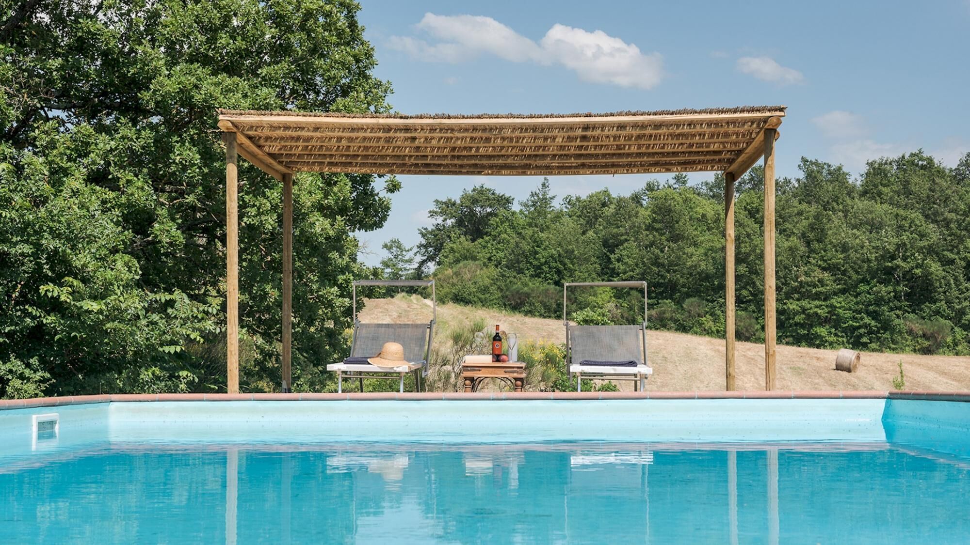 A poolside area with a pergola, two lounge chairs, and a table with a bottle and glasses.