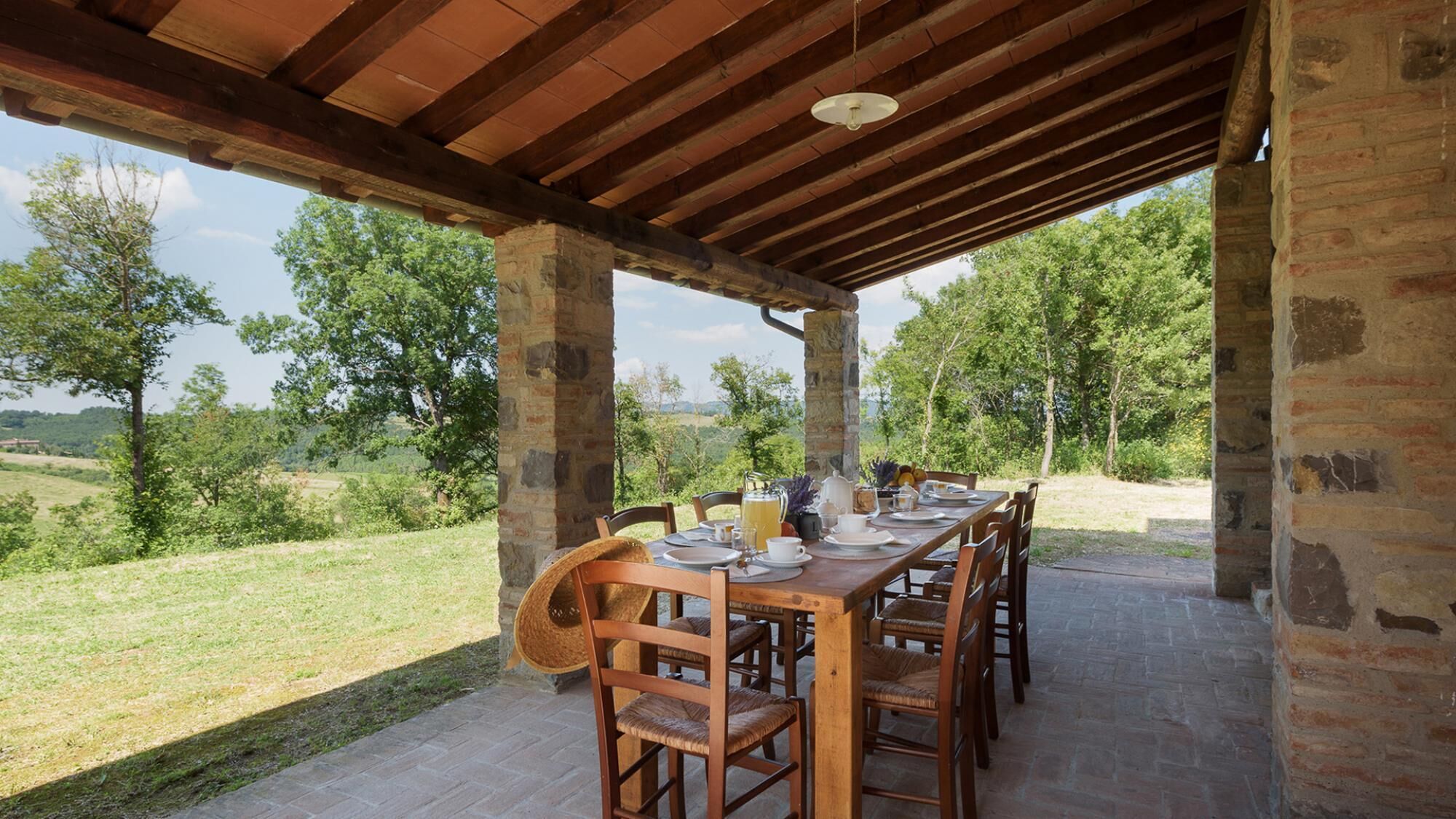 An outdoor dining area with a rustic wooden table and chairs, set against a backdrop of lush greenery and a clear sky.