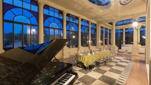 Een statige zaal met een piano, een lange eettafel gedekt voor een maaltijd en grote ramen met uitzicht op de lucht.