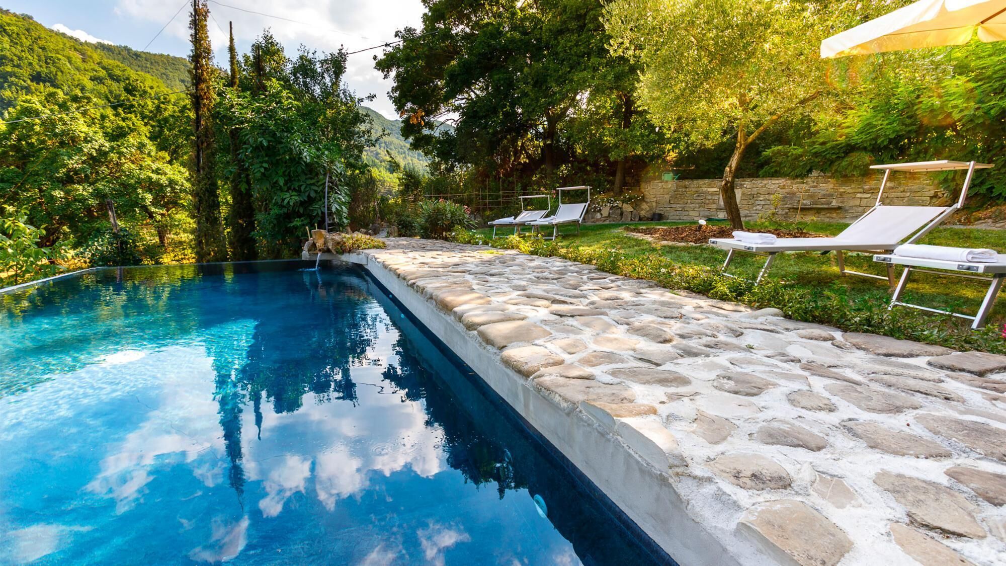 A swimming pool with stone steps leading into it, surrounded by greenery and a stone wall.