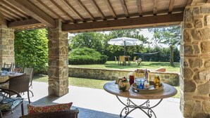 An outdoor patio with stone pillars, a round table with drinks, and a view of a grassy yard with a dining table and chairs. - Ar-d649-foch0ar - Villa Joy 8 (Foiano della Chiana)