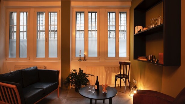 A cozy living room with a black leather sofa, a round coffee table, and a red armchair. There are large windows with frosted glass, a radiator, and a bookshelf with various items. - Apartment with sauna at the foot of the Citadel (Besançon)