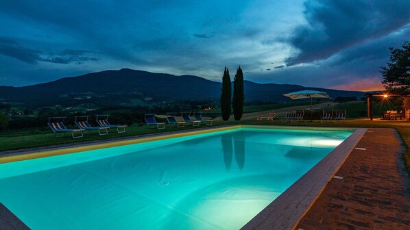 A swimming pool with lounge chairs and a pergola, surrounded by a brick pathway and a view of mountains.