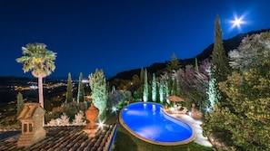 A nighttime view of a swimming pool surrounded by trees and a mountain backdrop.