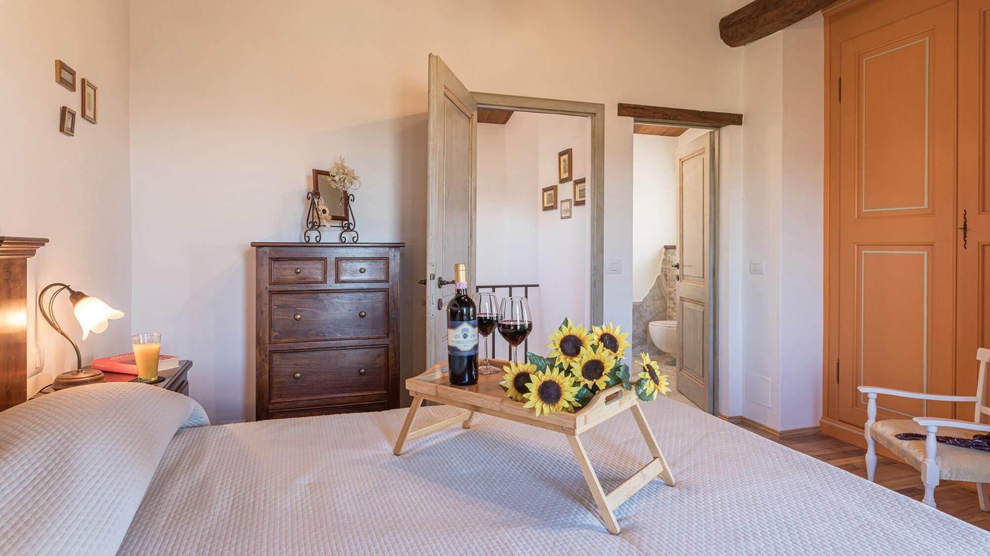 A bedroom with a bed, a dresser, a nightstand, a lamp, a mirror, a wooden stand with a wine bottle and glasses, and a chair.