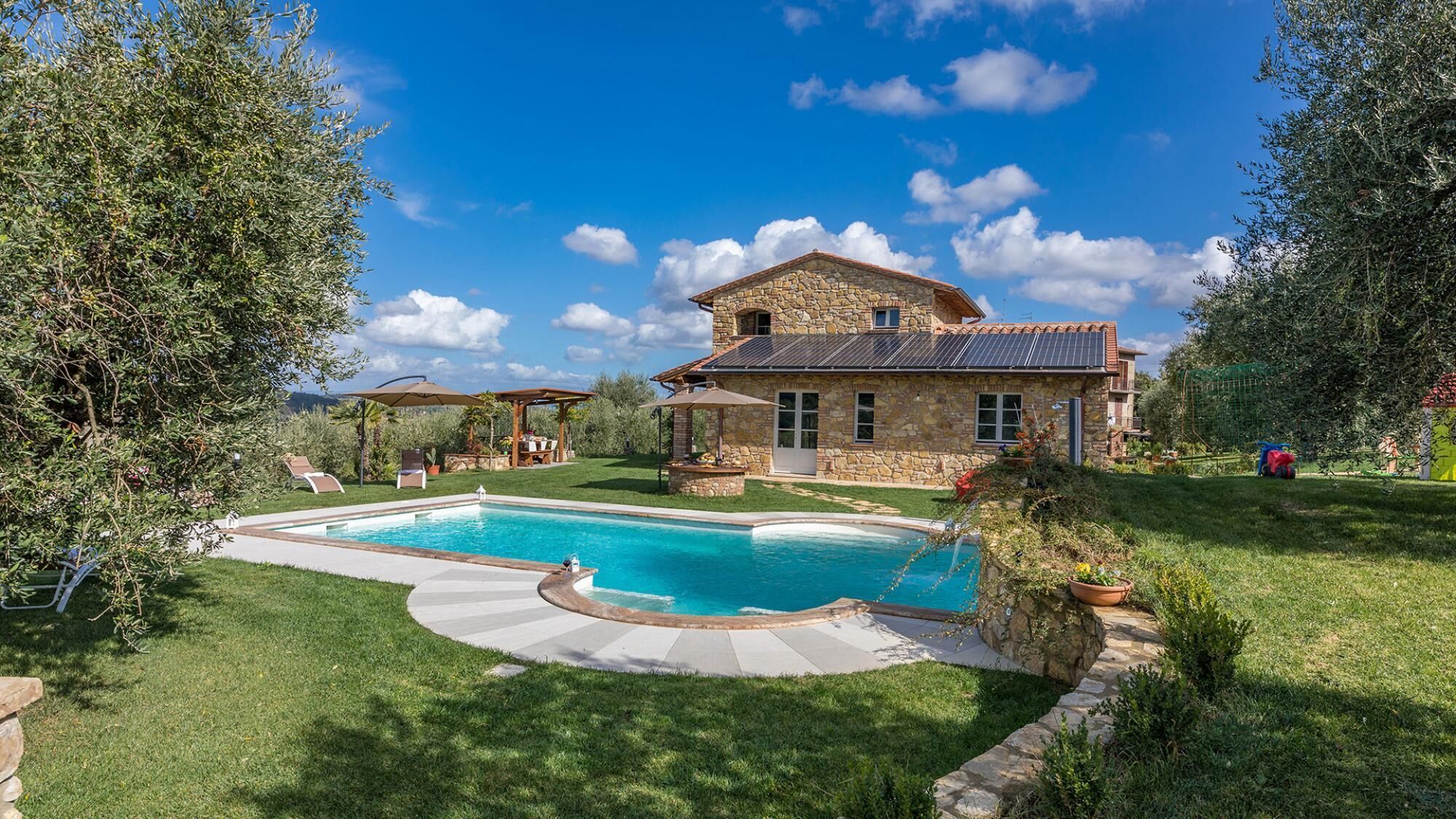 Una casa de piedra con piscina, rodeada por un césped cuidado y árboles.