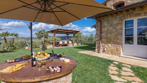A wooden outdoor dining area with a table set for a meal, a pergola with a table underneath, and a stone building with a glass door in the background. - Pg-c744-cipi0at - La Villetta di Dante 6 (Citta della Pieve)