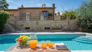 A poolside setting with a stone building, a fruit bowl, a pitcher of orange juice, and a book titled "The Guardians".