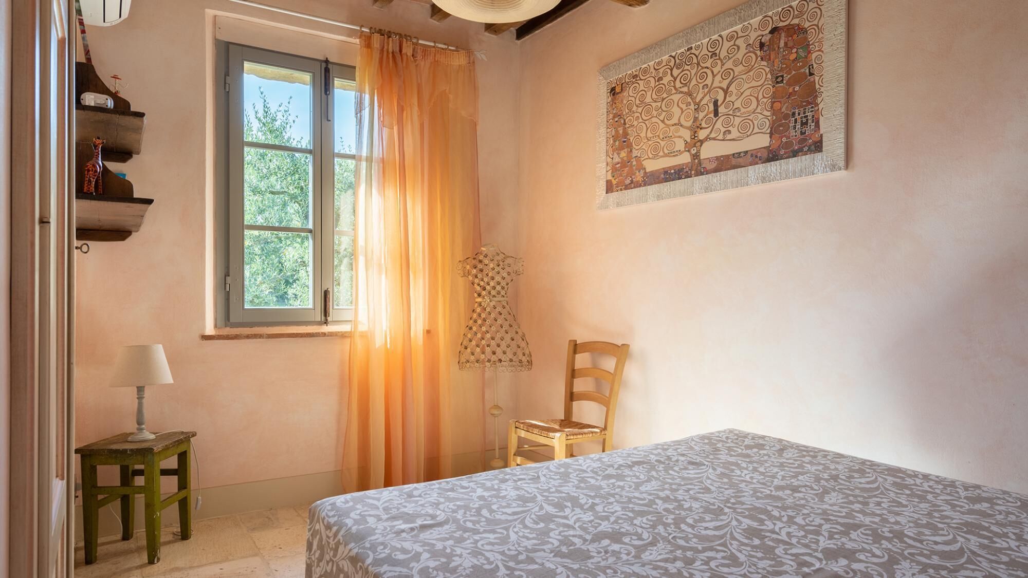 A bedroom with a bed, a chair, a lamp, a window with curtains, and a wall painting.