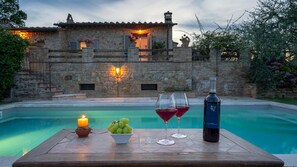 A stone building with a pool, a table with a candle, grapes, and wine glasses.