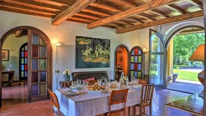 Un comedor interior con una mesa lista para una comida, vigas de madera en el techo y vistas del exterior a través de ventanas arqueadas.