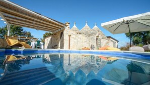 Ein Pool mit klarem Wasser, das die Umgebung spiegelt, ein Steinhaus mit traditionellem Dach und Sitzgelegenheiten im Freien mit Sonnenschirmen.