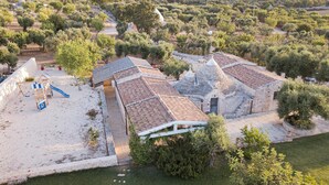 Um edifício de pedra tradicional com telhado de telhas, rodeado por oliveiras e uma área arenosa com um parquinho.