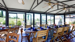 A covered outdoor dining area with tables set for breakfast, large windows offering views of the surrounding landscape, and ceiling fans. - Gr-d948-lpog0at - Villa Croci 26 (Gavorrano)