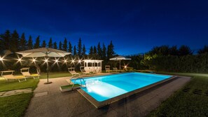 Une piscine rectangulaire, bordée d’une terrasse carrelée, entourée de chaises longues et de parasols, le tout se fondant dans un décor de verdure et sous un ciel dégagé.