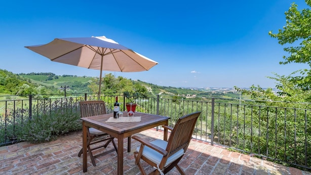 A patio with a table and chairs, a large umbrella, and a scenic view of rolling hills. - An-f453-cbgn2at - Casale Verdicchio (Montecarotto)