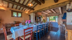 A dining area with a long table set for a meal, wooden beams on the ceiling, and a brick fireplace. - An-f453-cbgn2at - Casale Verdicchio (Montecarotto)