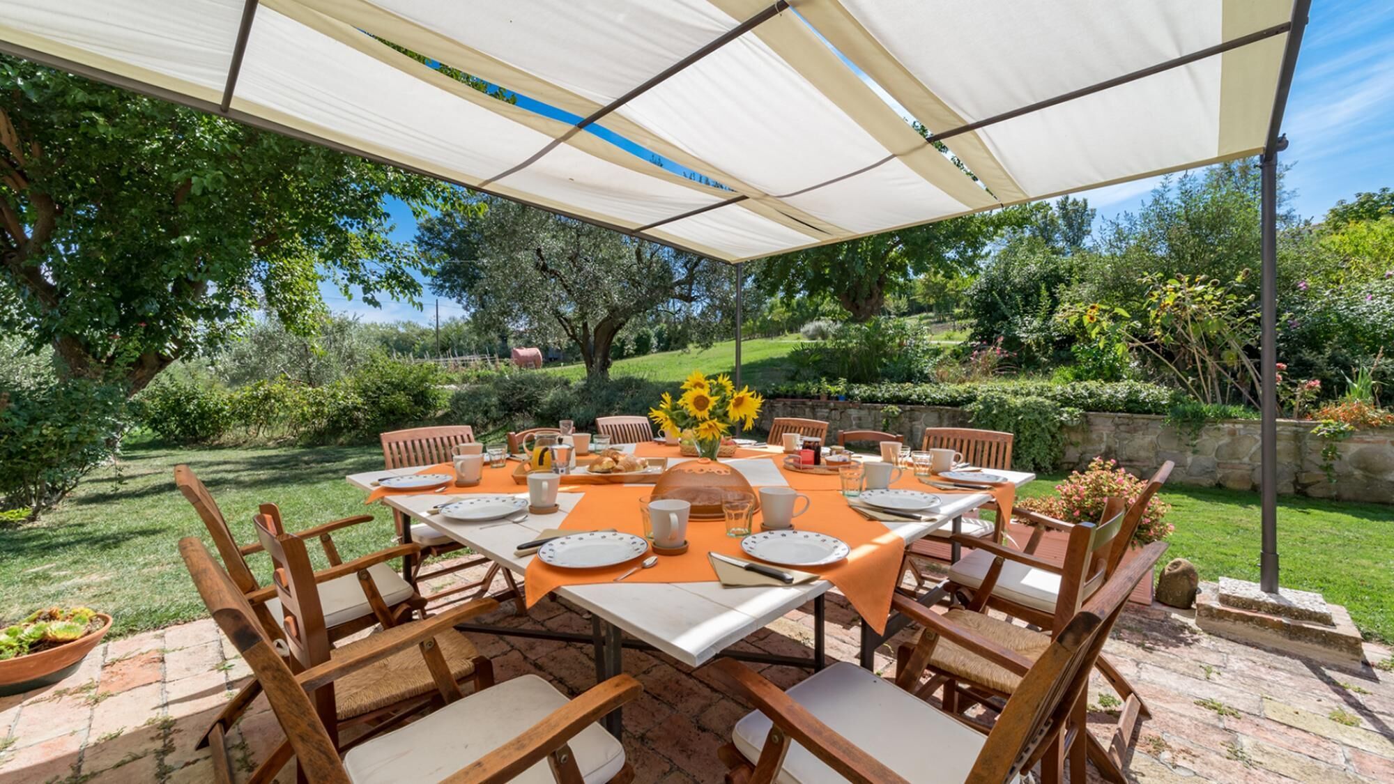 Zona d'exterior per dinar amb una taula preparada per a vuit persones, amb caires de fusta, sota un gran toldo.