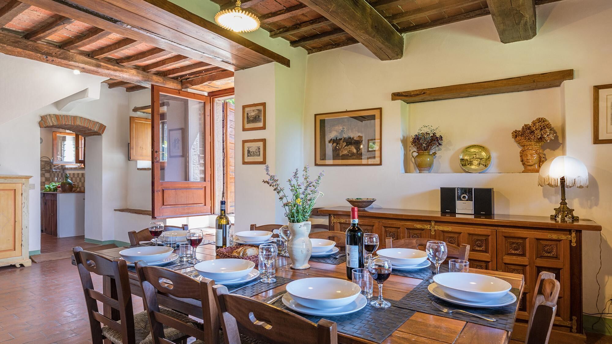 Een eethoek met een houten tafel voor vier personen, omringd door stoelen. Er is een keuken zichtbaar door een geopende deur, en een eettafel met een bloevas en flessen wijn.