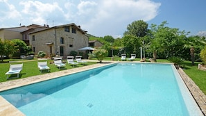 A rectangular swimming pool with clear blue water, surrounded by a stone border. There are several white lounge chairs with blue cushions arranged around the pool. The pool area is adjacent to a stone building with a tiled roof and multiple windows. - Si-i877-sp3714at - La Capanna di Franca 6 2 (Sovicille)