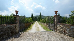 A gravel path leading to a gated entrance with stone walls and metal gates, flanked by greenery and trees. - Si-i877-sp3714at - La Capanna di Franca 6 2 (Sovicille)