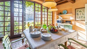 Un comedor con una mesa preparada para el desayuno, vistas al exterior a través de grandes ventanales y un estante con utensilios de cocina.