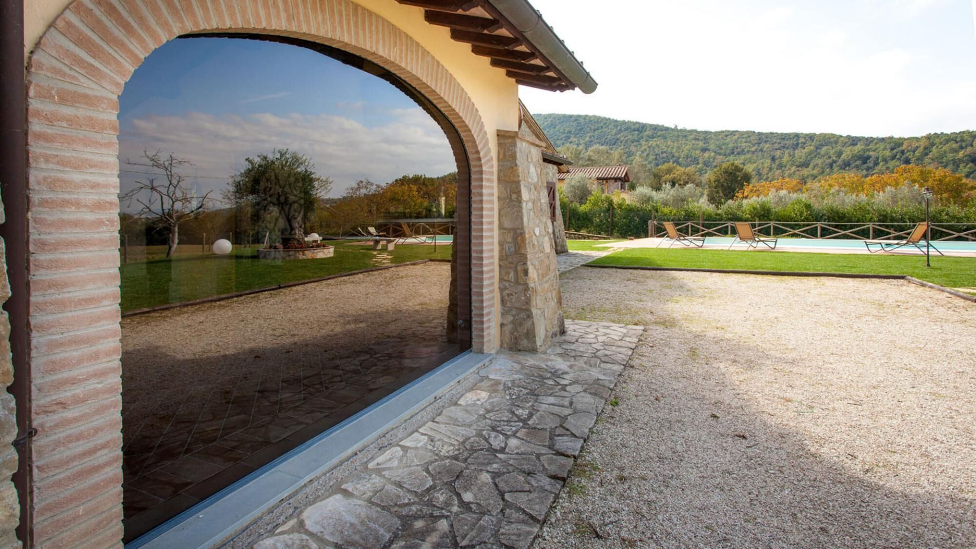 Un arco de piedra que da acceso a un patio con piscina, rodeado por un sendero de grava y un césped.