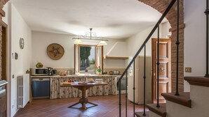 A kitchen with a dining table, a hanging light fixture, a view through the window, and a staircase leading to another level. - Si-a468-spla0at - Fattoria L Olmo Rosso 14 (Sinalunga)