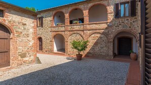 A courtyard with a gravel ground, a potted tree, and a building with stone walls and arched windows. - Si-a468-spla0at - Fattoria L Olmo Rosso 14 (Sinalunga)