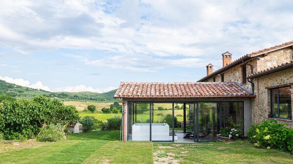 A stone house with a tiled roof, a glass-enclosed patio with a hot tub, and a lush green garden.