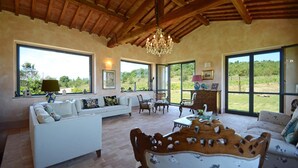 A spacious living room with a white sofa, wooden chairs, a coffee table, and a chandelier. Large windows offer views of the outdoors. - Tr-i381-svim0at - Podere Pomasciano 10 (San Venanzo)