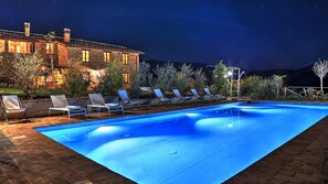Une piscine avec des chaises longues, un bâtiment en briques illuminé et un ciel nocturne.