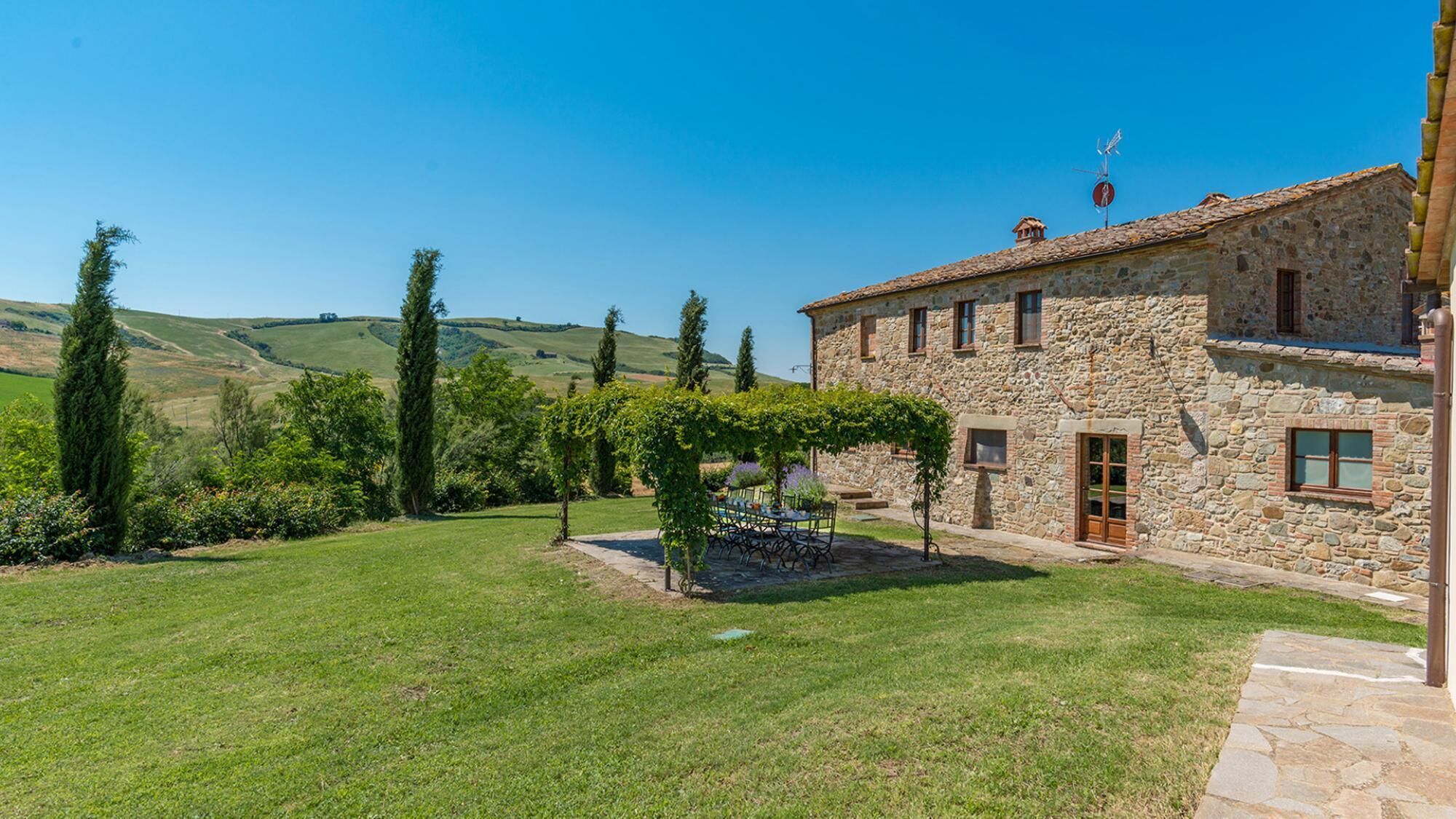A stone house with a stone roof, a pergola with a table and chairs, and a clear view of rolling hills in the background.