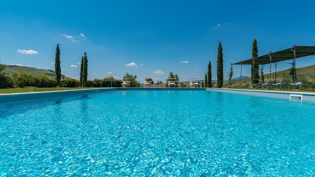 Una piscina cristallina con sedie a sdraio e ombrelloni sul lato destro, immersa nel verde e con dolci colline sullo sfondo.