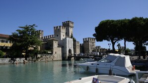 A historic castle with a prominent tower, situated by a calm body of water with boats docked nearby. - Bs-i633-viem71a1 - Arcimboldo House 2 2 (Sirmione)