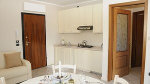 A compact kitchen with white cabinets, a stove, and a sink. There is a dining table set with plates, glasses, and cutlery. A wooden door and a white wall with a telephone are visible. - Bs-i633-viem71a1 - Arcimboldo House 2 2 (Sirmione)