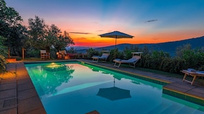A swimming pool with lounge chairs and an umbrella, surrounded by greenery and hills in the background.