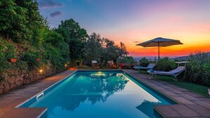 Piscina con sillas de descanso y una sombrilla, rodeada de follaje y vista al atardecer.